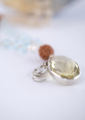 A beaded necklace with light blue, clear, and brown beads draped over a large blue crystal geode, with a bright yellow rose prop in the foreground on a white background.