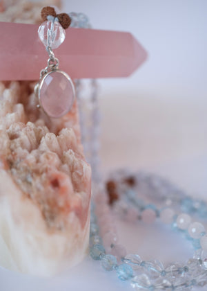 A delicate beaded necklace with light blue, pale pink, and brown beads, featuring a large oval pink stone pendant and a small silver crescent moon charm, displayed on a clear heart-shaped tray next to a pink geode.