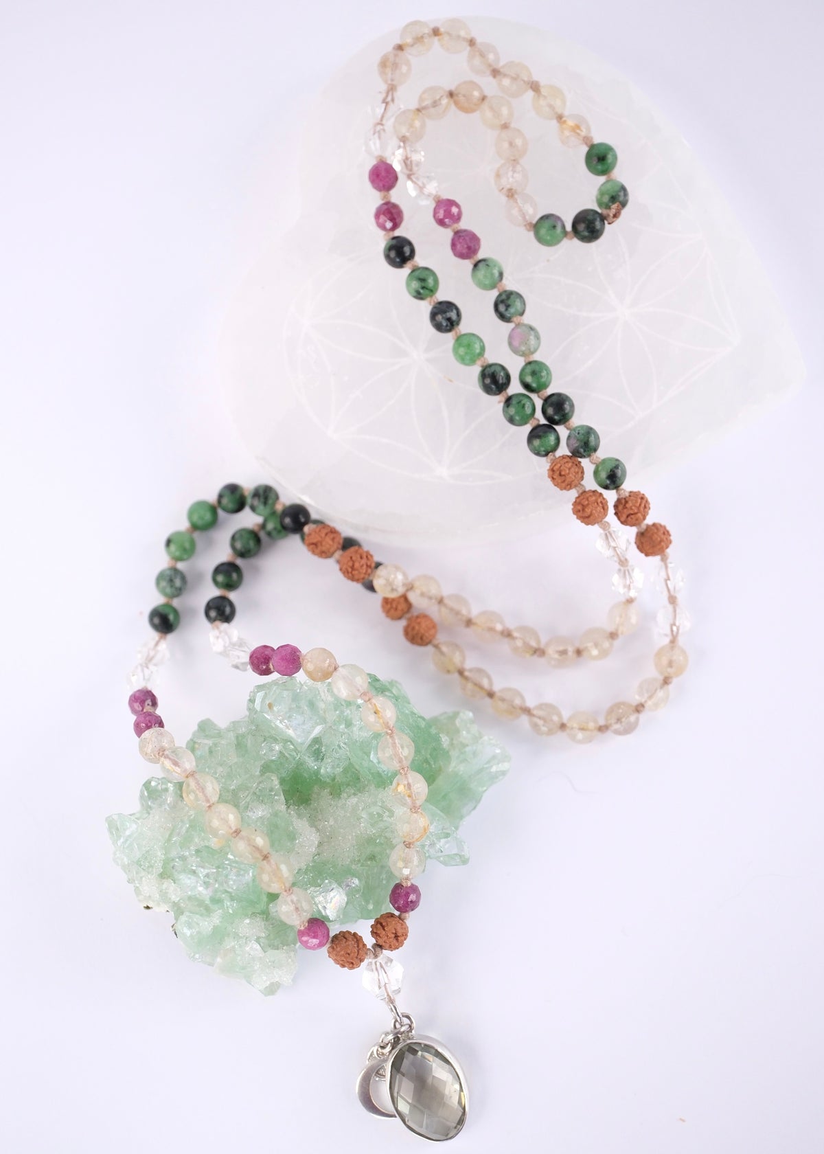 A beaded necklace with green, black, pink, and tan beads, featuring a light green crystal cluster and a silver-toned heart pendant, displayed on a white background with a white geometric prop.