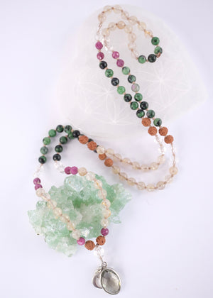 A beaded necklace with green, black, pink, and tan beads, featuring a light green crystal cluster and a silver-toned heart pendant, displayed on a white background with a white geometric prop.