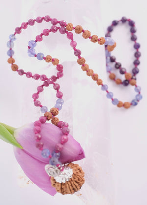 A hand holding a beaded necklace featuring a large pink ruby, faceted purple amethyst beads, rudraksha, ruby and tanzanite