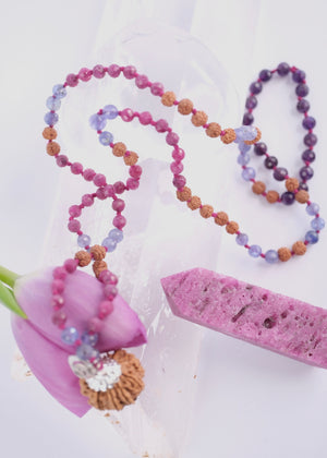 A hand holding a beaded necklace featuring a large pink ruby, faceted purple amethyst beads, rudraksha, ruby and tanzanite
