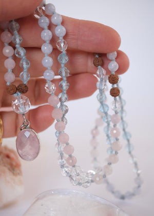 A delicate beaded necklace with light blue, pale pink, and brown beads, featuring a large oval pink stone pendant and a small silver crescent moon charm, displayed on a clear heart-shaped tray next to a pink geode.