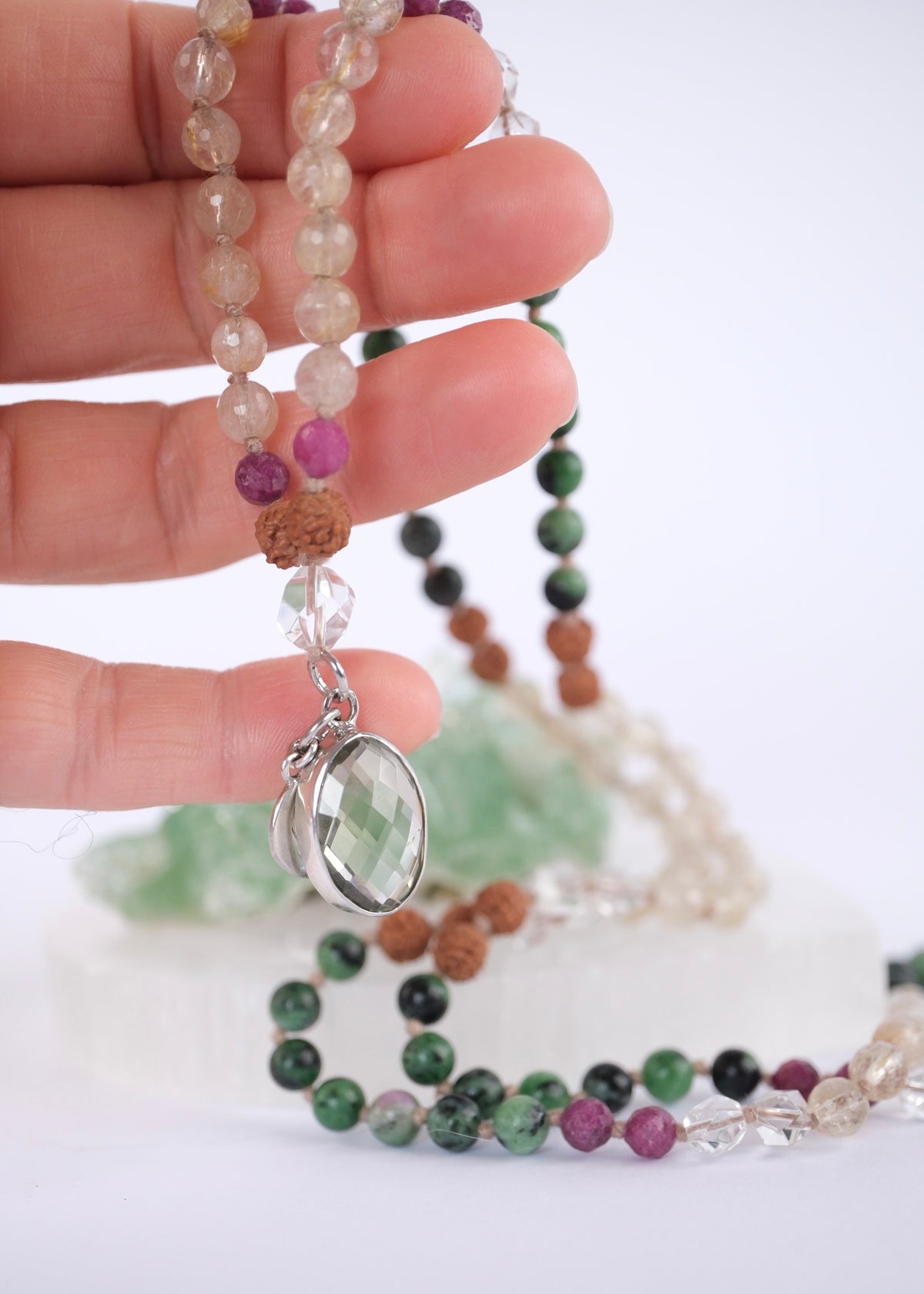 A close-up of a hand holding a beaded necklace with a faceted green crystal pendant, alongside other strands of green, brown, purple, and clear beads on a white background.