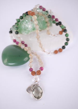 A beaded necklace with green, black, pink, and tan beads, featuring a light green crystal cluster and a silver-toned heart pendant, displayed on a white background with a white geometric prop.