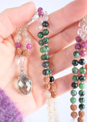 A beaded necklace with green, black, pink, and tan beads, featuring a light green crystal cluster and a silver-toned heart pendant, displayed on a white background with a white geometric prop.