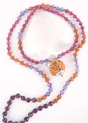 A hand holding a beaded necklace featuring a large pink ruby, faceted purple amethyst beads, rudraksha, ruby and tanzanite