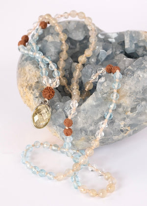 A beaded necklace with light blue, clear, and brown beads draped over a large blue crystal geode, with a bright yellow rose prop in the foreground on a white background.