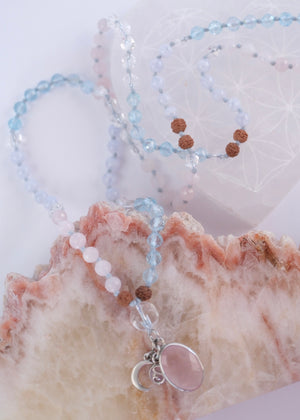 A delicate beaded necklace with light blue, pale pink, and brown beads, featuring a large oval pink stone pendant and a small silver crescent moon charm, displayed on a clear heart-shaped tray next to a pink geode.