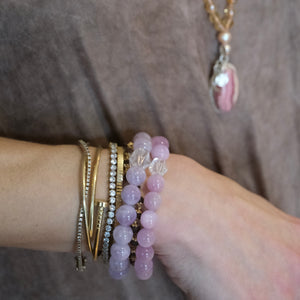 A close-up of a person's wrist wearing a stack of bracelets, including pink beaded bracelets, gold bangles, and a silver rhinestone bangle, against a brown textured background.