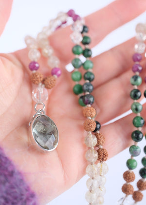 A beaded necklace with green, black, pink, and tan beads, featuring a light green crystal cluster and a silver-toned heart pendant, displayed on a white background with a white geometric prop.