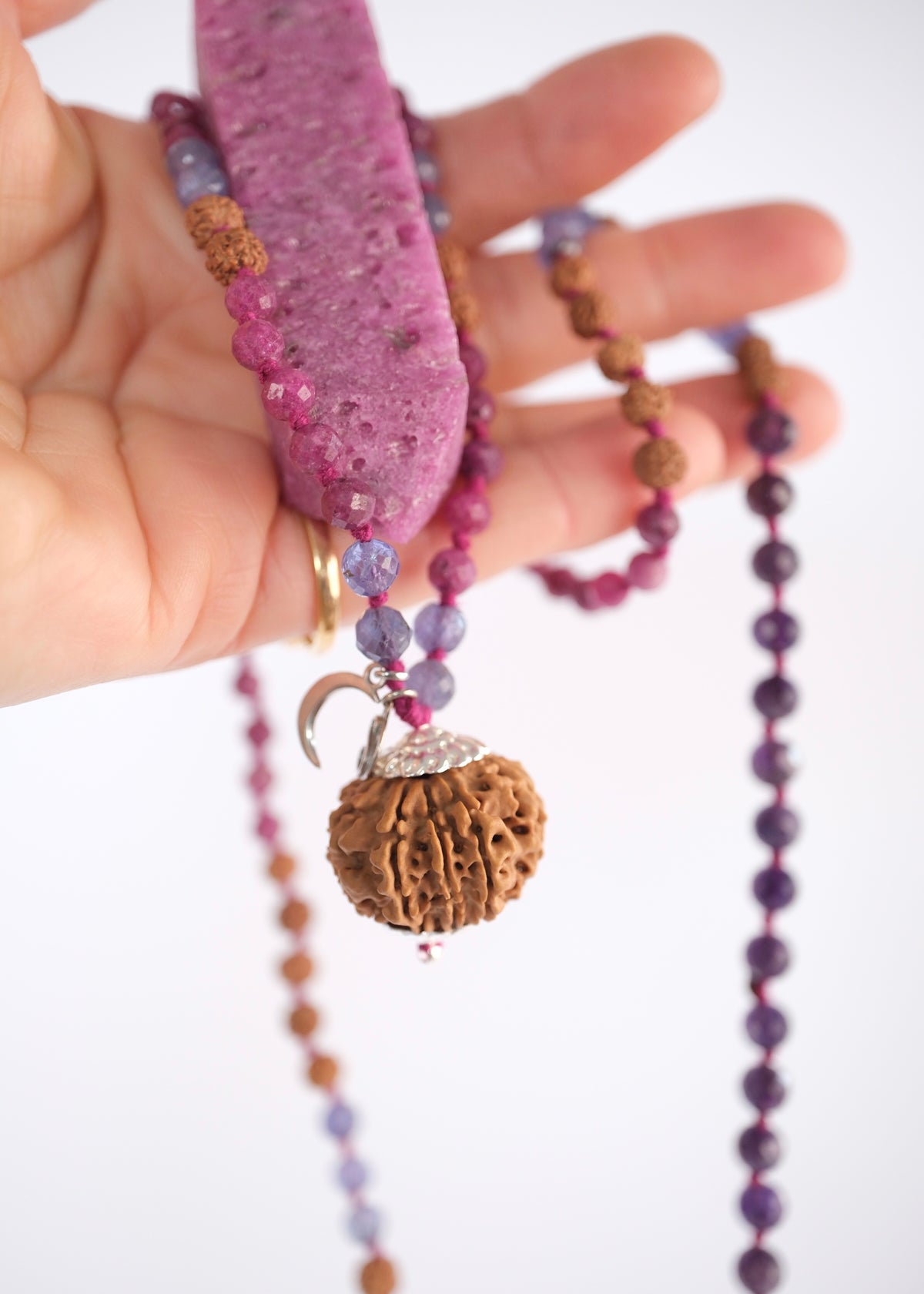 A hand holding a beaded necklace featuring a large pink ruby, faceted purple amethyst beads, rudraksha, ruby and tanzanite