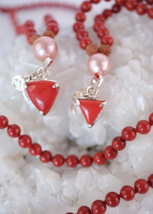 A red beaded necklace with pink pearl-like beads, brown textured spacers, and two dangling red triangular charms with silver-toned metal details, photographed on a white background.