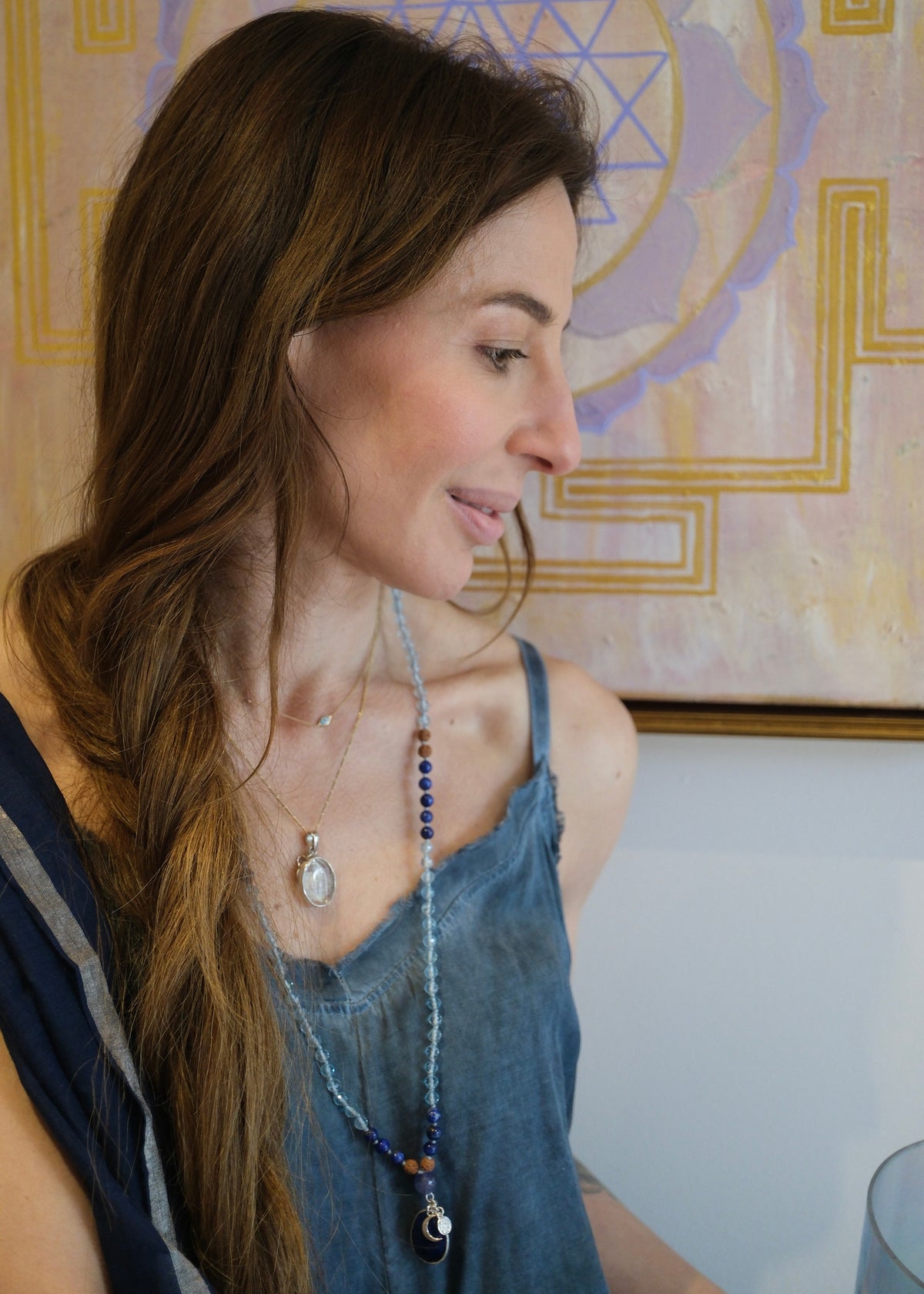 A woman with long brown hair wearing a blue denim top and layered necklaces, including a long beaded necklace with a dark pendant, photographed indoors in front of a decorative wall art piece.