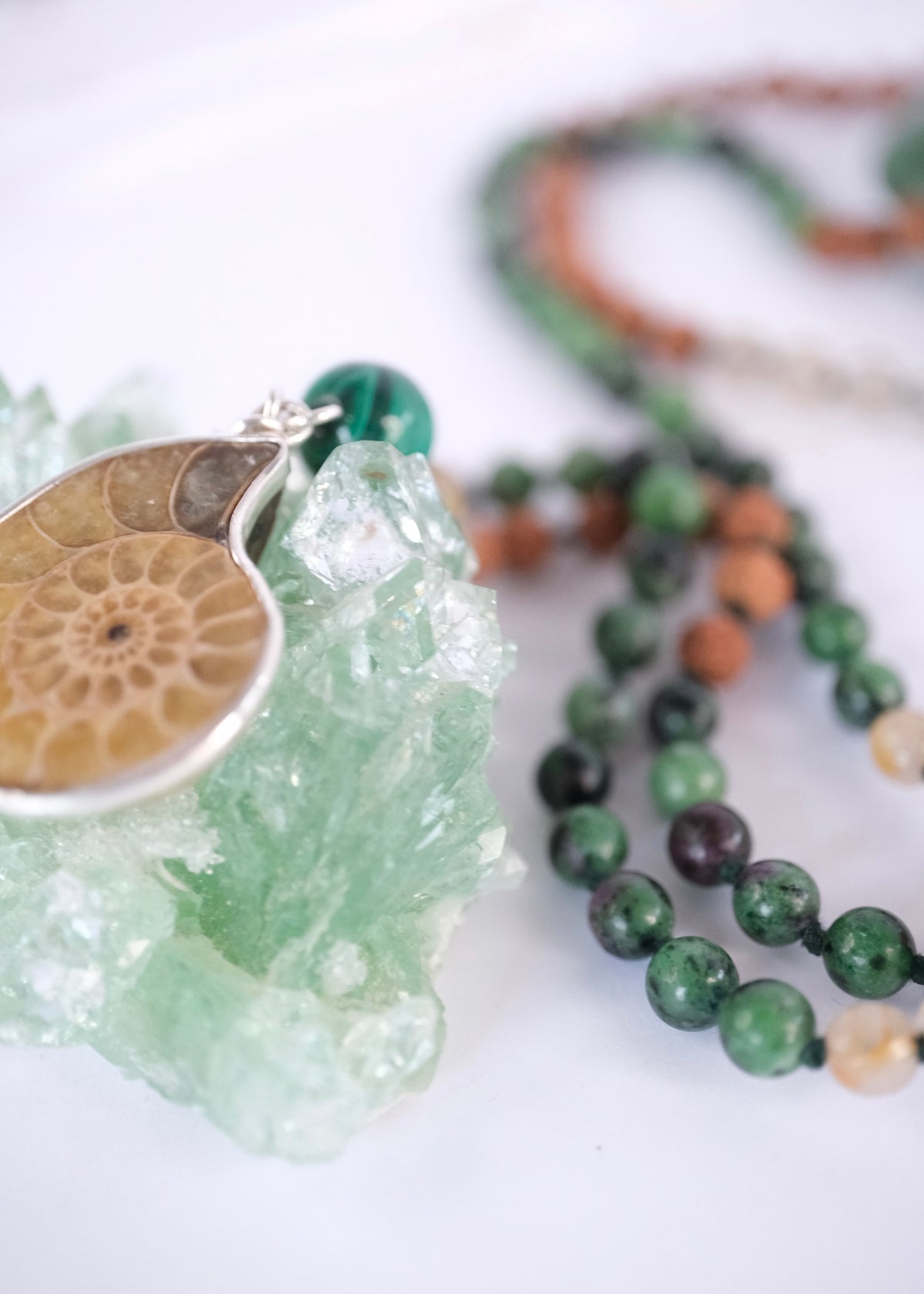 A close-up of a beaded necklace with green, brown, and cream beads, featuring a silver-toned pendant shaped like a fossilized ammonite, displayed on a light green crystal cluster against a white background.