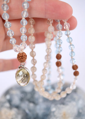 A beaded necklace with light blue, clear, and brown beads draped over a large blue crystal geode, with a bright yellow rose prop in the foreground on a white background.