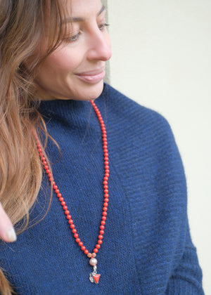 A red beaded necklace with pink pearl-like beads, brown textured spacers, and two dangling red triangular charms with silver-toned metal details, photographed on a white background.
