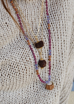 A hand holding a beaded necklace featuring a large pink ruby, faceted purple amethyst beads, rudraksha, ruby and tanzanite