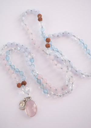 A delicate beaded necklace with light blue, pale pink, and brown beads, featuring a large oval pink stone pendant and a small silver crescent moon charm, displayed on a clear heart-shaped tray next to a pink geode.
