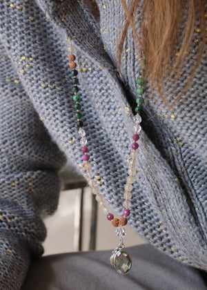 A beaded necklace with green, black, pink, and tan beads, featuring a light green crystal cluster and a silver-toned heart pendant, displayed on a white background with a white geometric prop.