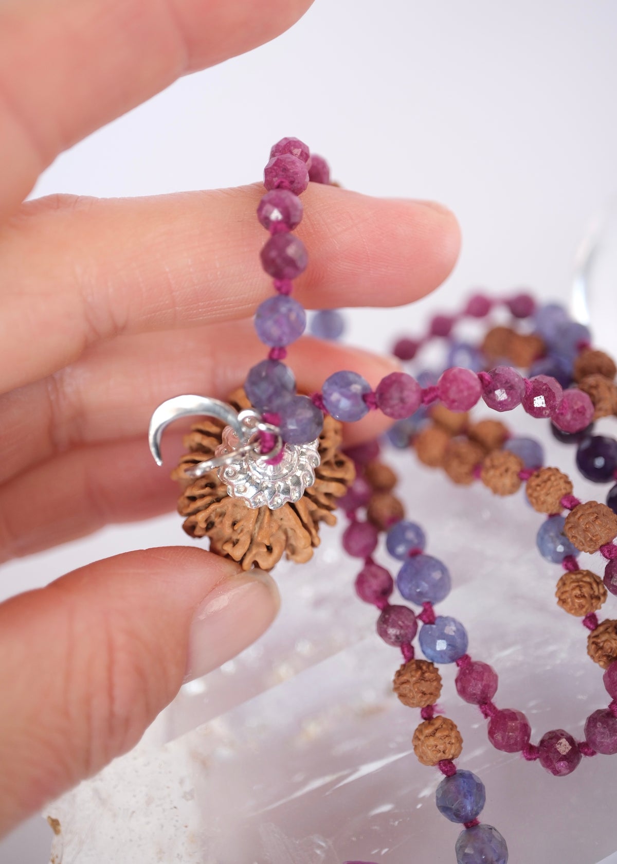 A close-up of a hand holding a beaded necklace with faceted purple and blue beads, brown textured beads, a silver crescent moon, and a silver sunburst charm with a clear crystal center.