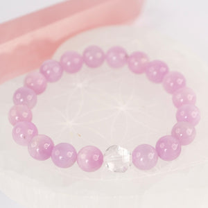 A close-up of a person's wrist wearing a stack of bracelets, including pink beaded bracelets, gold bangles, and a silver rhinestone bangle, against a brown textured background.