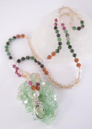 A beaded necklace with green, black, pink, and tan beads, featuring a light green crystal cluster and a silver-toned heart pendant, displayed on a white background with a white geometric prop.