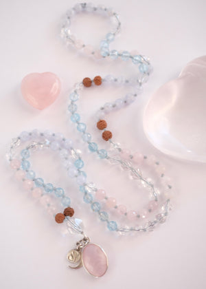 A delicate beaded necklace with light blue, pale pink, and brown beads, featuring a large oval pink stone pendant and a small silver crescent moon charm, displayed on a clear heart-shaped tray next to a pink geode.