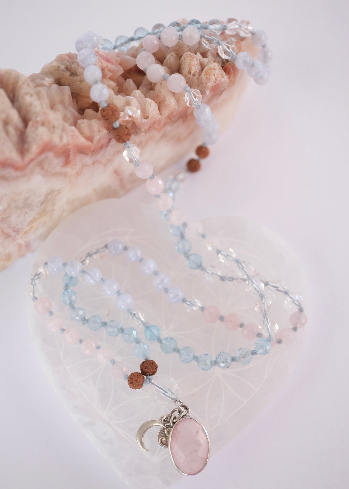 A delicate beaded necklace with light blue, pale pink, and brown beads, featuring a large oval pink stone pendant and a small silver crescent moon charm, displayed on a clear heart-shaped tray next to a pink geode.