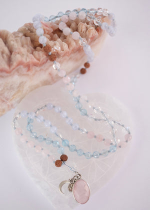 A delicate beaded necklace with light blue, pale pink, and brown beads, featuring a large oval pink stone pendant and a small silver crescent moon charm, displayed on a clear heart-shaped tray next to a pink geode.