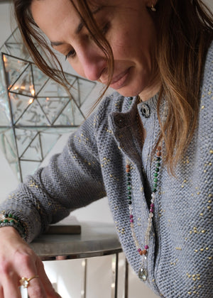 A beaded necklace with green, black, pink, and tan beads, featuring a light green crystal cluster and a silver-toned heart pendant, displayed on a white background with a white geometric prop.