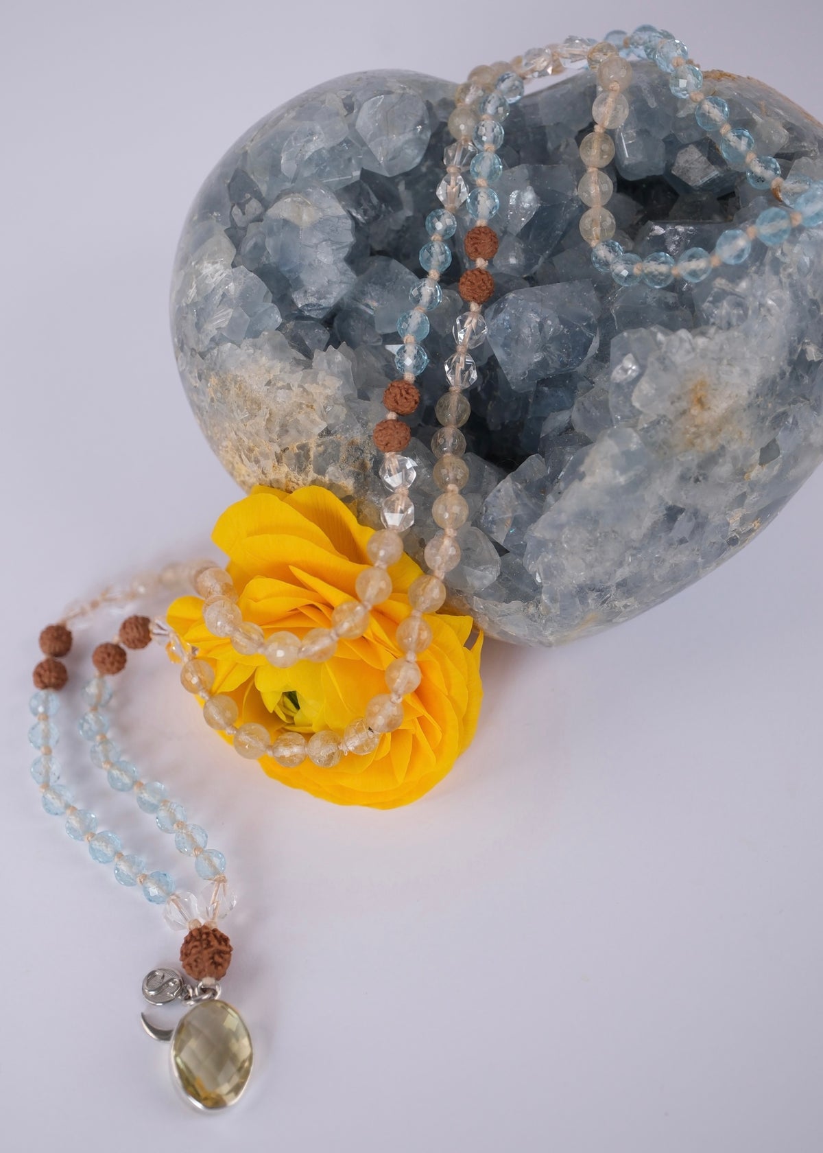 A beaded necklace with light blue, clear, and brown beads draped over a large blue crystal geode, with a bright yellow rose prop in the foreground on a white background.
