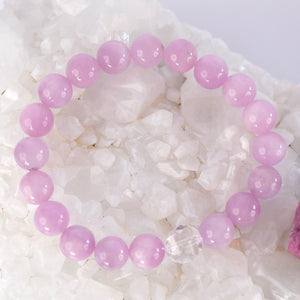 A close-up of a person's wrist wearing a stack of bracelets, including pink beaded bracelets, gold bangles, and a silver rhinestone bangle, against a brown textured background.