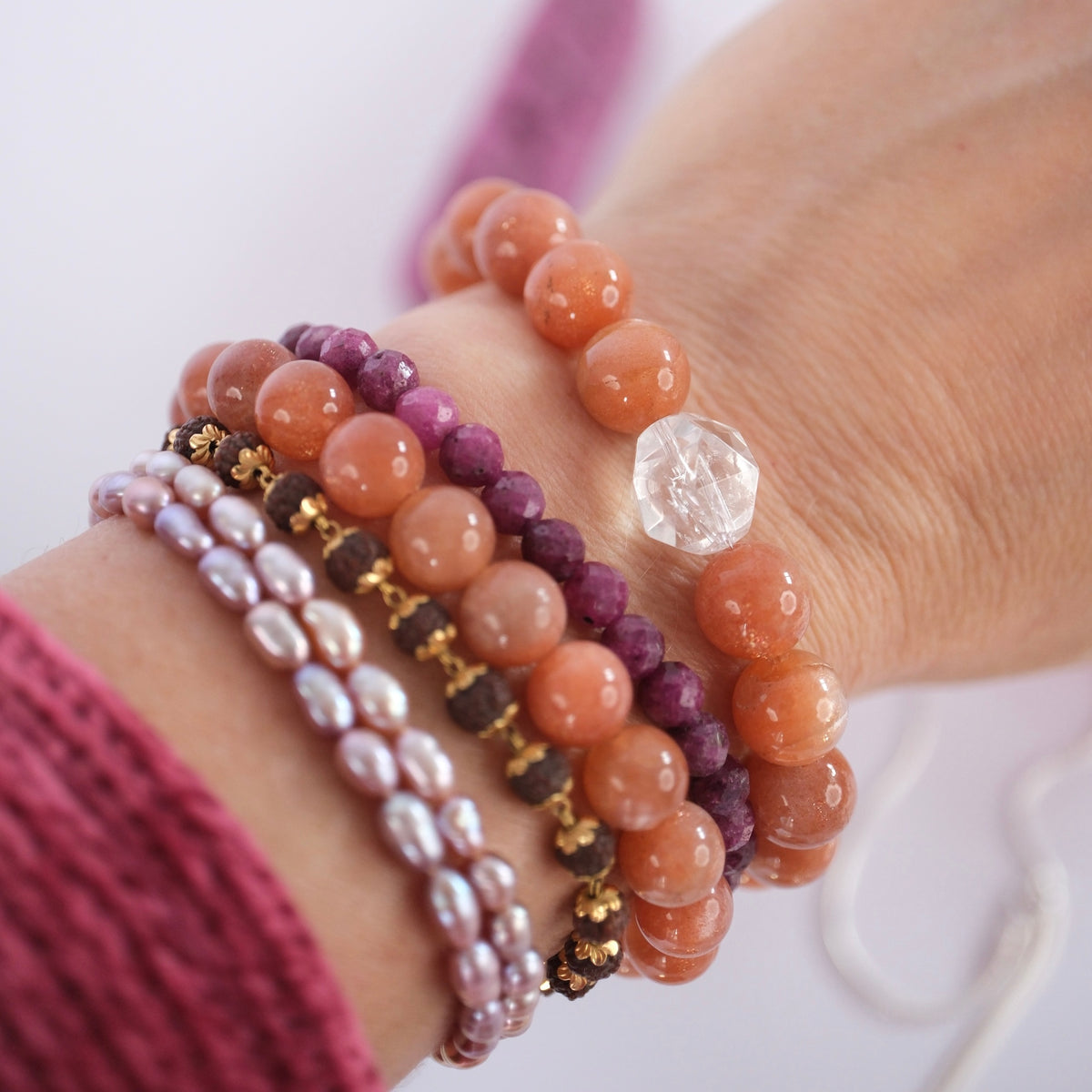 A close-up of a person's wrist wearing multiple beaded bracelets. The bracelets include orange stone beads, purple stone beads, white pearl-like beads with gold accents, dark brown beads, and a clear faceted bead.