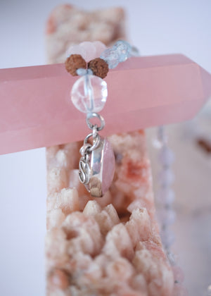 A delicate beaded necklace with light blue, pale pink, and brown beads, featuring a large oval pink stone pendant and a small silver crescent moon charm, displayed on a clear heart-shaped tray next to a pink geode.